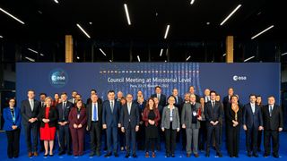 several dozen well-dressed people stand in front of a blue backdrop at a conference