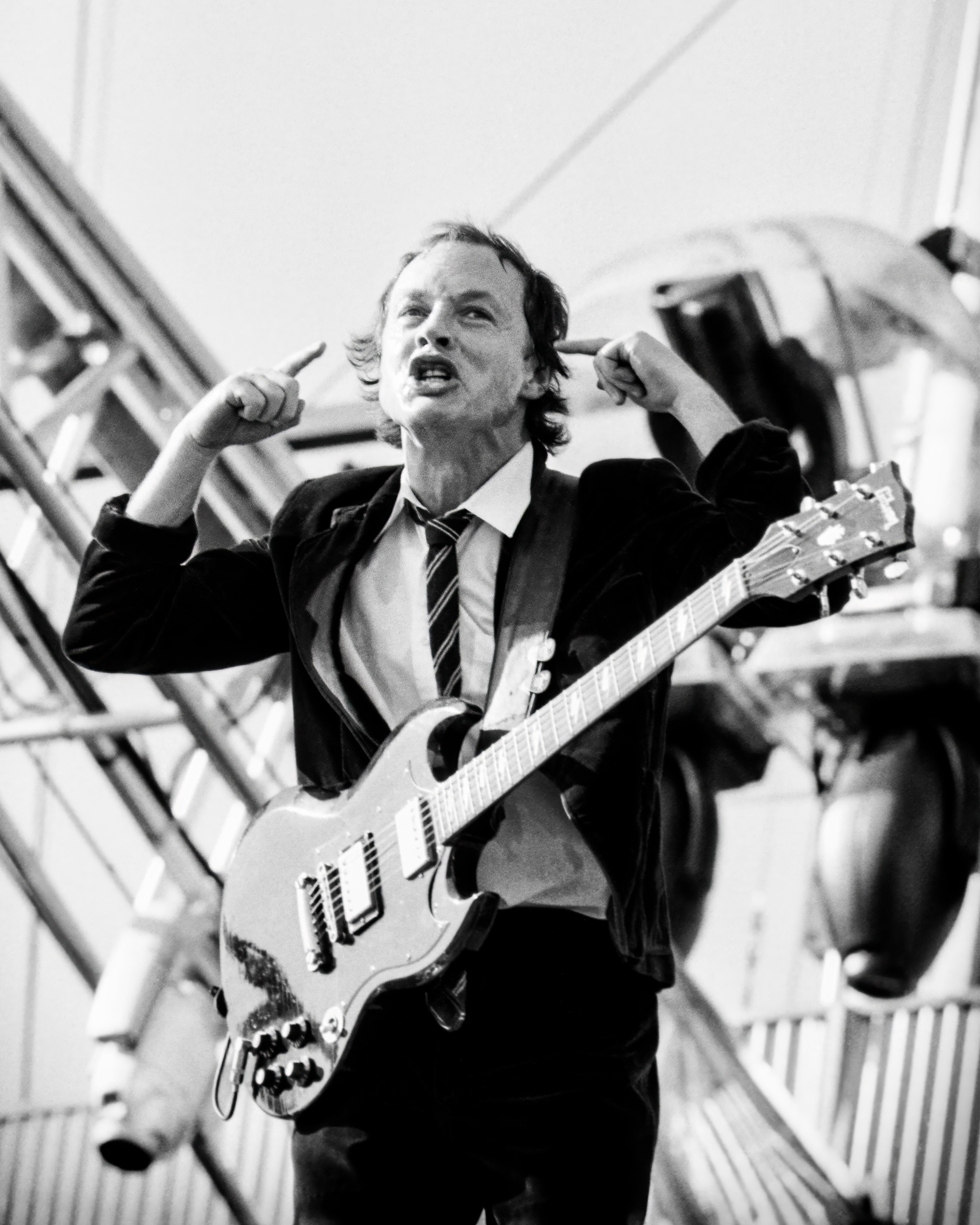 Angus Young performs in black and white with fingers raised beside his head and mouth open mid-shout, stage scaffolding visible behind him.