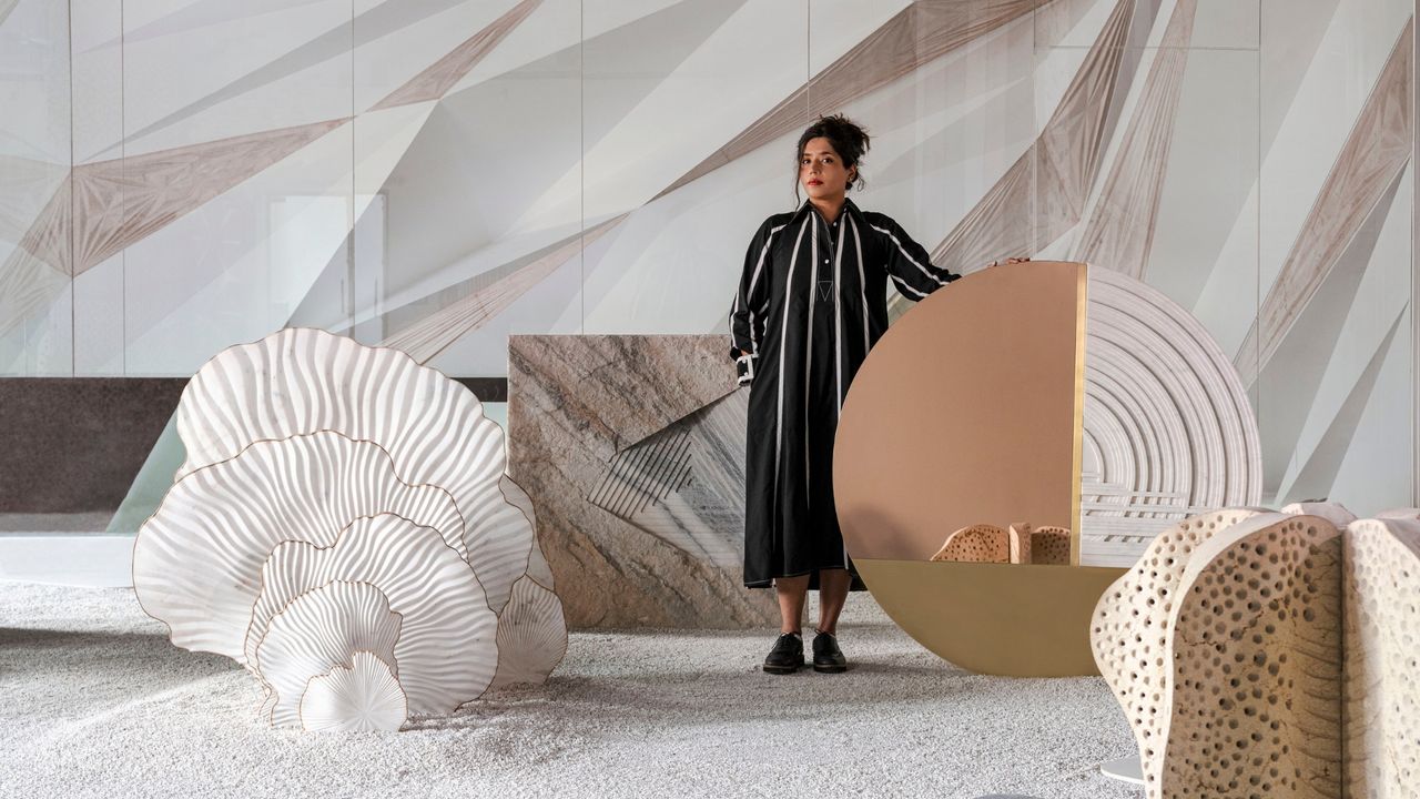 A young woman dressed in a stripy black and white maxi dress stands in a neutral-colored room filled with architectural installations on the tones of sand, cream, brown, and white.