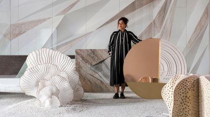 A young woman dressed in a stripy black and white maxi dress stands in a neutral-colored room filled with architectural installations on the tones of sand, cream, brown, and white.