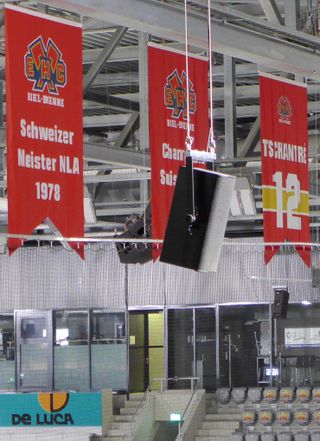 Silver Alcons Audio speakers hang from the rafters in front of several red banners at an ice hockey arena.