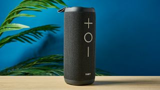 a black oblong-shaped bluetooth speaker with mesh covering and a carry strap photographed on a beige table in front of a navy wall