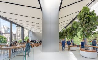 Alternative interior view of the upper level at Apple's San Francisco store featuring light coloured flooring, floor-to-ceiling windows, long wooden tables, stools, trees in round planters and a large stainless steel central structure. There are multiple people in the store