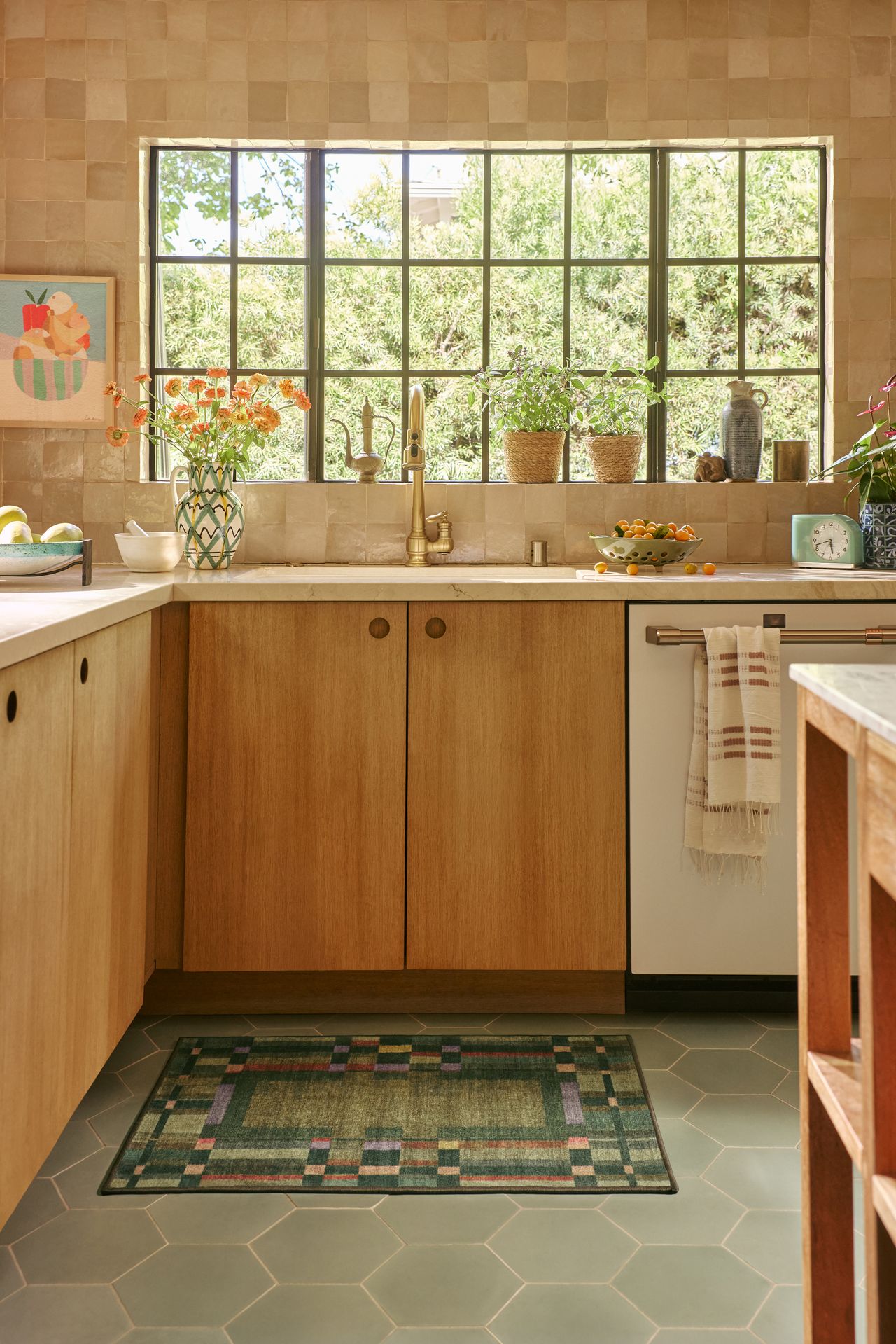 Image of a contemporary kitchen with wooden cabinetry, sage green tile floors, and tan tile walls. There is a green, geometric rug under the sink.