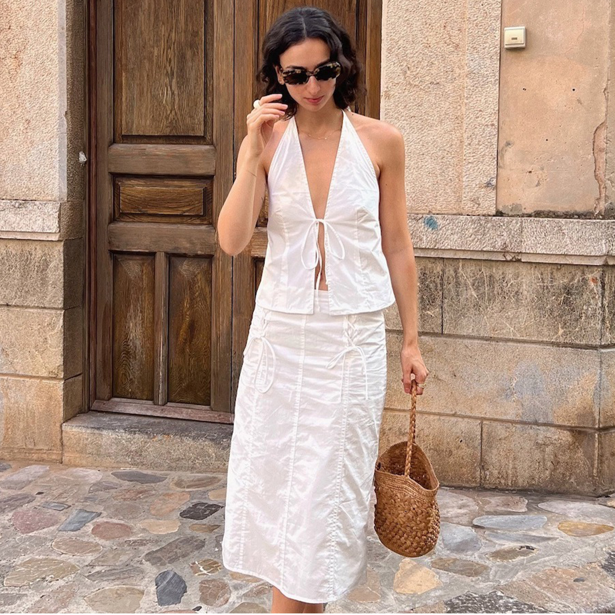 A woman wearing a white linen skirt and top holding a woven brown bucket bag. 