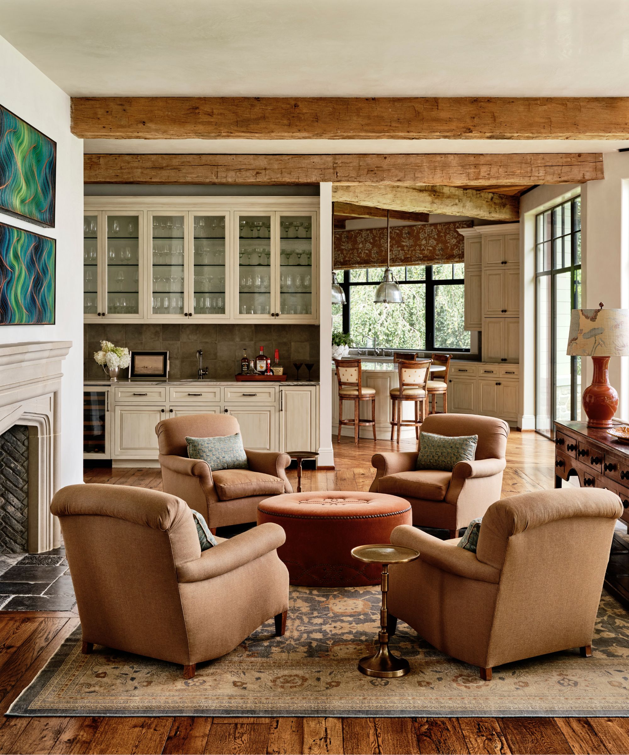 cozy fall living room with wooden ceiling beams, a large rug with four chestnut colored arm chairs, a pumpkin colored ottoman and a large fireplace with views through to the rustic kitchen