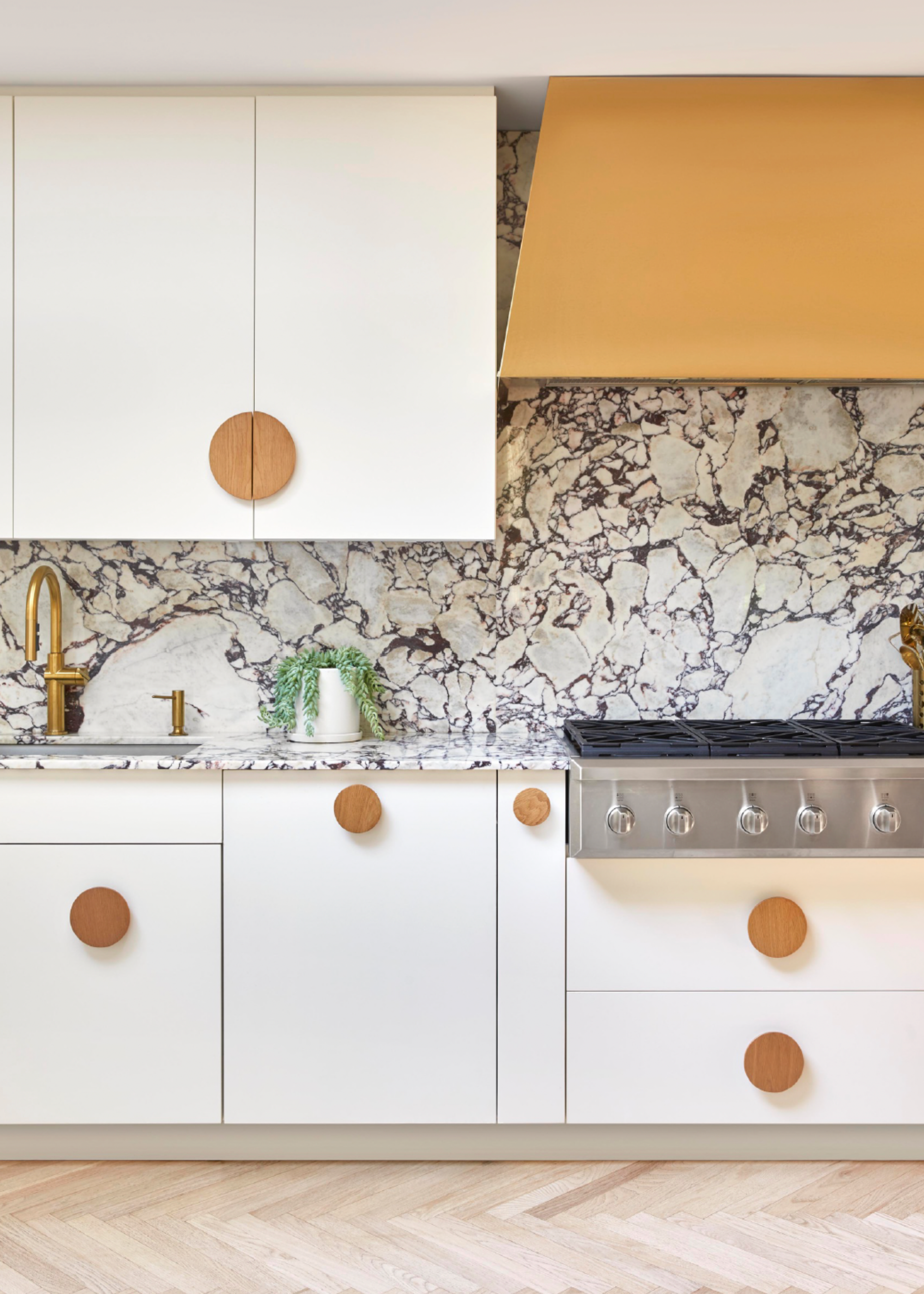 a cream kitchen with light herringbone flooring and marble countertop and backsplash, yellow extractor hood, and large round cabinet pulls