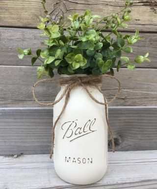 A white mason jar which says 'Ball mason' on it with green stems in it and twine around it, standing on a distressed gray wooden table