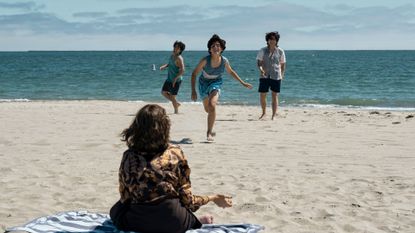 a woman (sofia vergara as griselda) sitting on a blue striped beach towel with her back to the camera faces three boys running towards her on the sand, with the sea in the background
