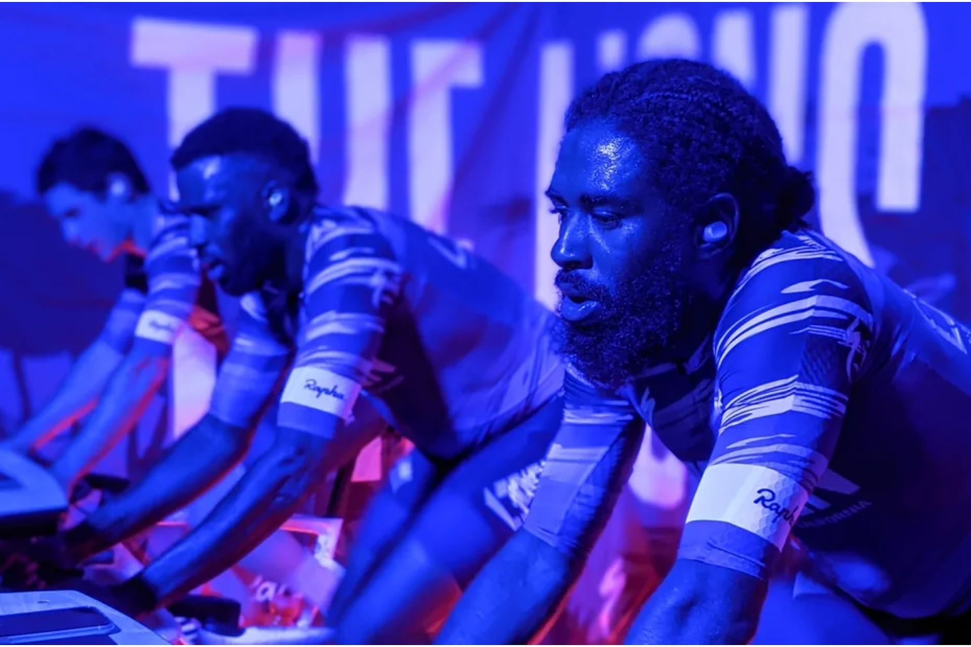 This image has a purple filter. Light. Jaybird Vista 2 are the best headphones for cycling as they are shown here on a rider on a static bike. You only see his head and torso. He is wearing a Rapha top, he is sweating, and there are other riders in the same kit in the background.