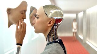 Alicia Vikander as the robot Ava in the movie Ex Machina touching a fake human face hanging on a white wall.