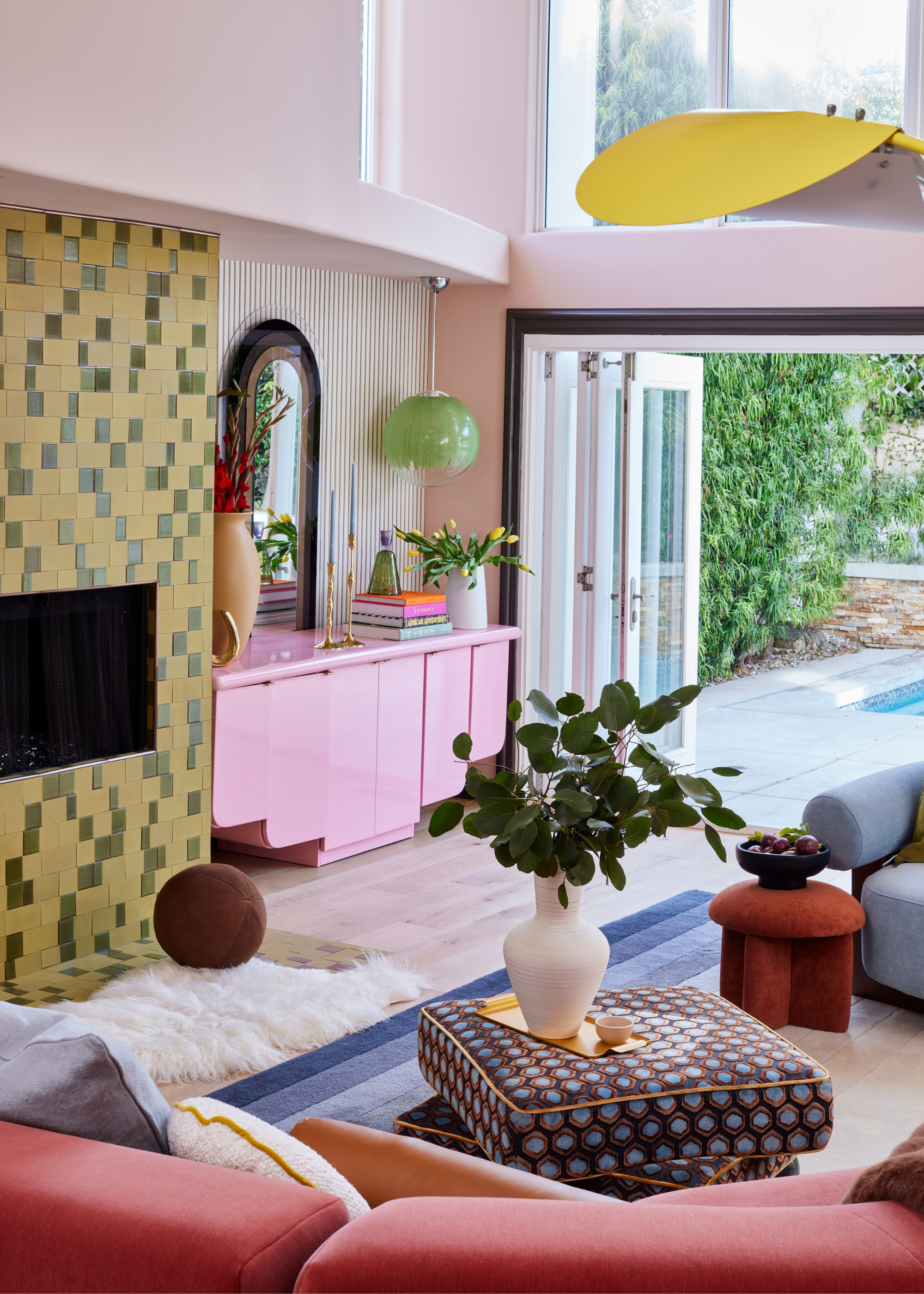An eclectic maximalist living room with a colorfully tiled fireplace, a pink sideboard, a patterned stacked coffee table, a shaggy floor covering, a mustard stool, and vases of stems