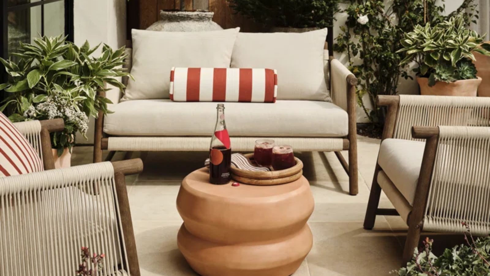 patio area surrounded by potted plants with wooden rope style patio furniture set with neutral seat cushions and a red striped bolster