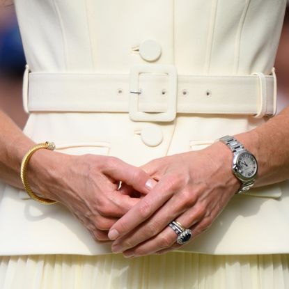 A closeup of Kate Middleton wearing a pale yellow dress, watch, bangle bracelet and stack of engagement/wedding rings