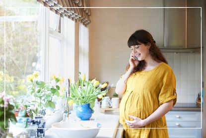 pregnant woman in yellow dress talking on phone while standing by the sink in the kitchen