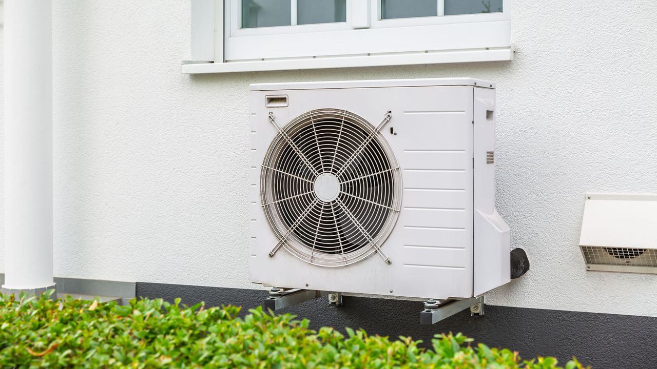 White heat pump on the side of a white house, above a green hedge