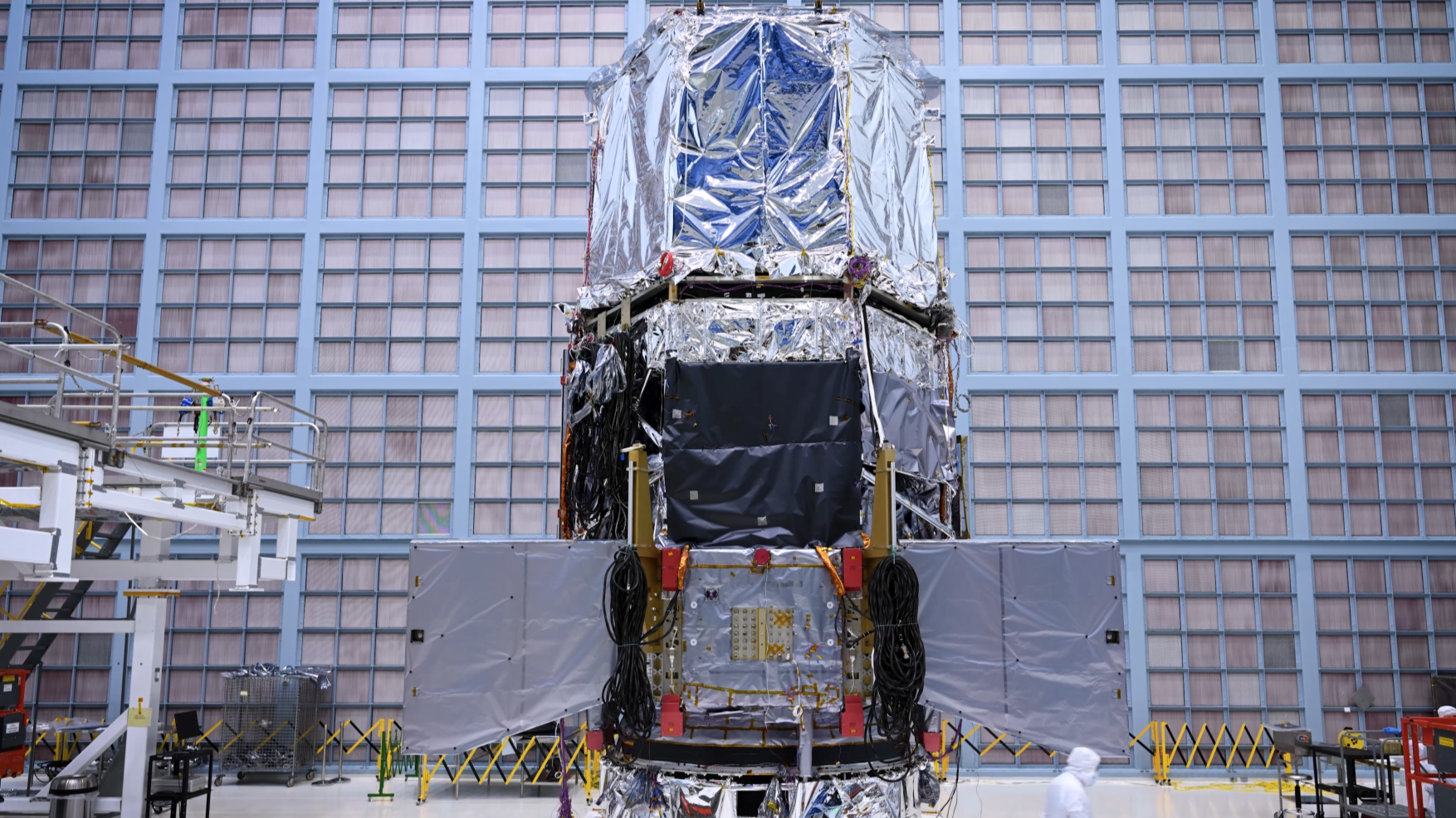 The Nancy Grace Roman Space Telescope, a large piece of covered equipment, displays part of its sunshield, a rolled up piece of white fabric, in its cleanroom