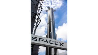 a giant shiny silver rocket stands behind a sign that says "spacex," under a cloudy blue skyu