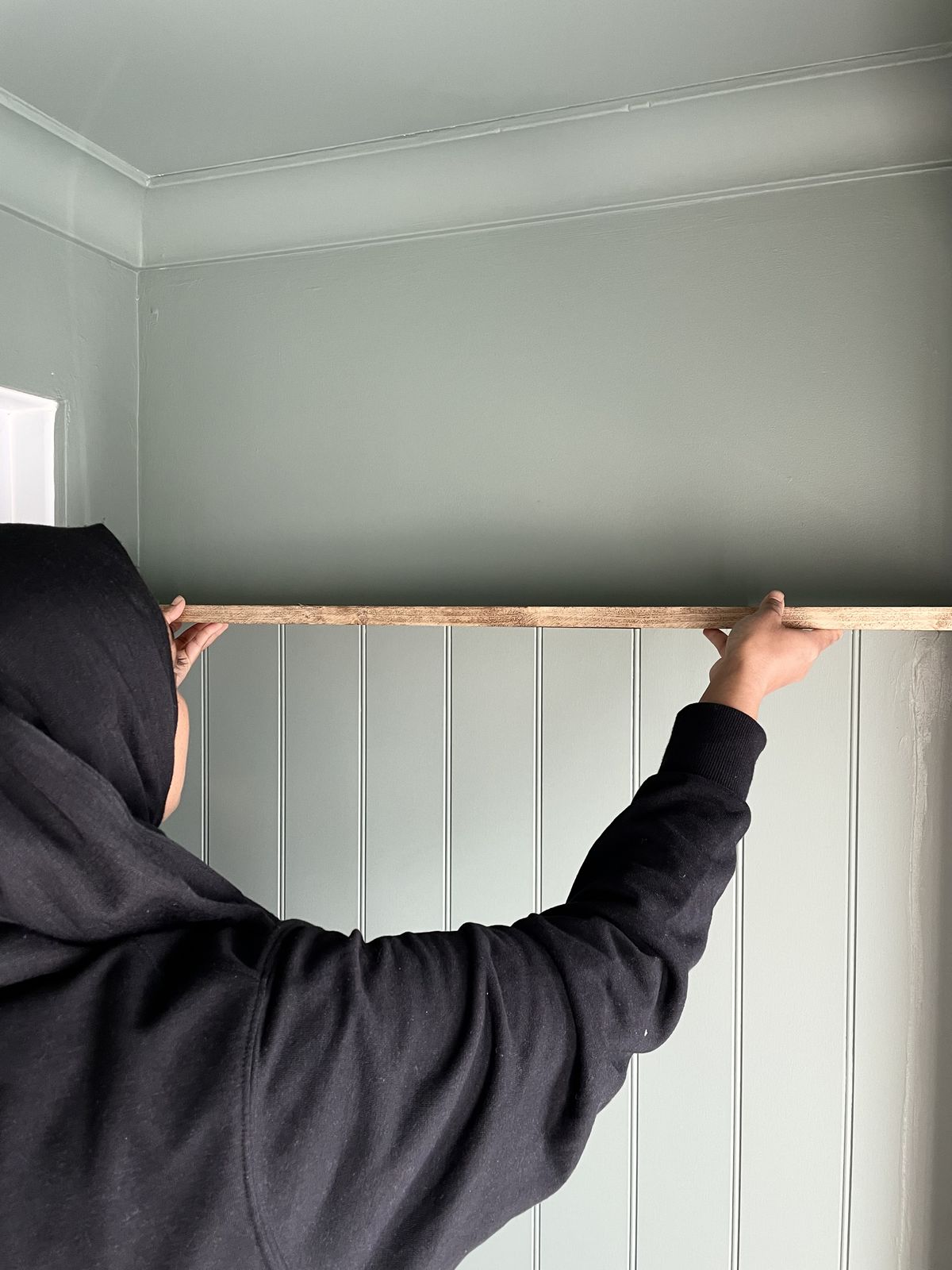 A woman positions a floating shelf into place above wood panelled walls