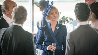 Jane Horrocks in a blue fascinator and suit holds a glass of champagne and talks to men in suits in COBRA: Rebellion.