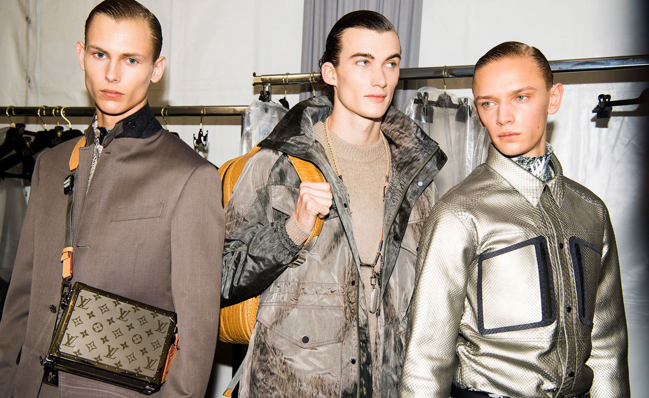 Three male models wearing looks from Louis Vuitton&#039;s collection. One model is wearing a grey jacket and bag with orange elements featuring the Louis Vuitton signature pattern. Next to him is a model wearing a beige jumper, long necklace, grey jacket and orange backpack. And the third model is wearing a light coloured metallic and blue shirt with a patterned high neck top underneath