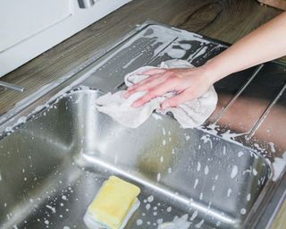 Arm of person cleaning grey stainless steel sink with lots of soapy water and a sponge