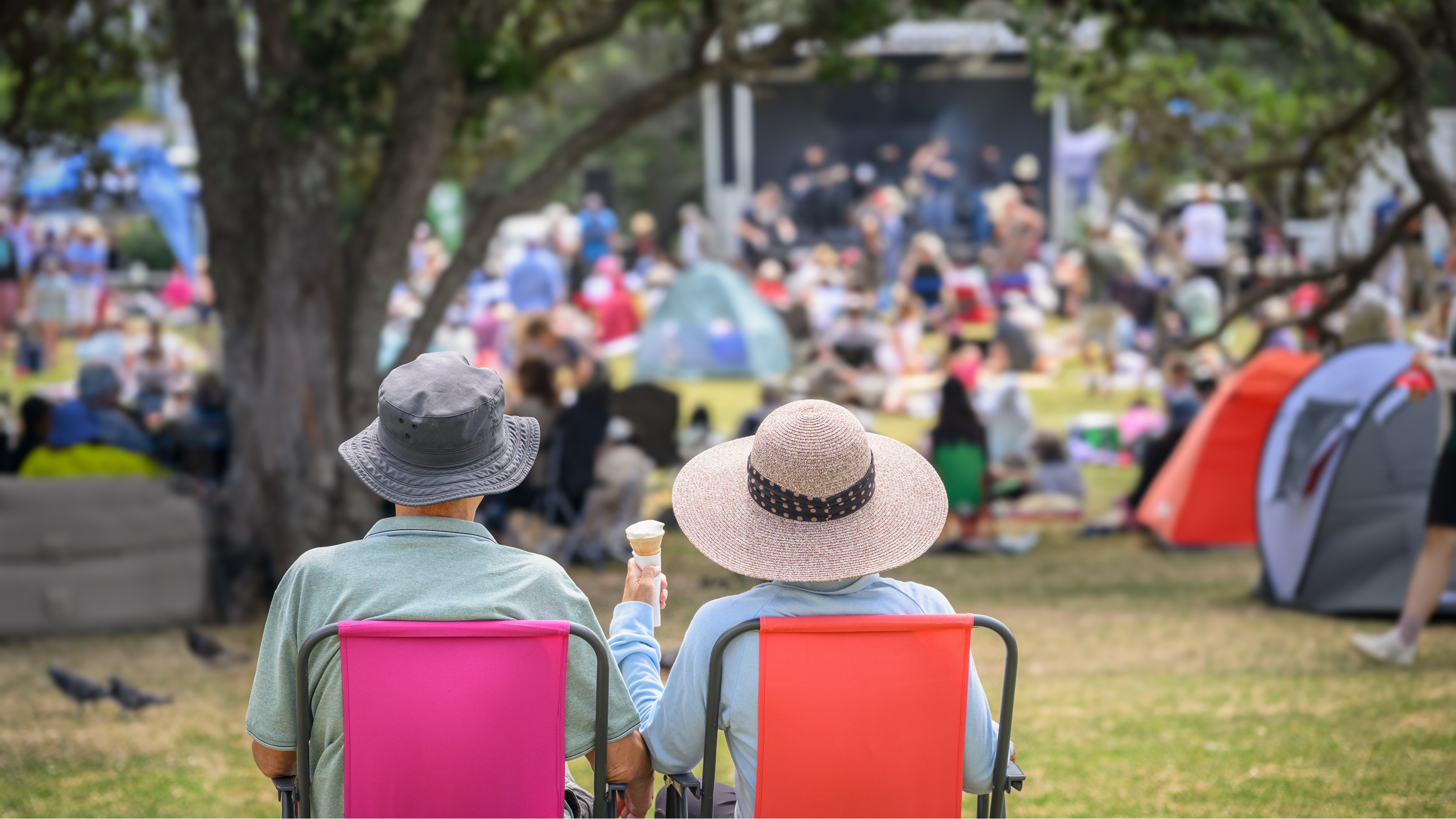 An older couple in lawn chairs enjoy some music in the park.