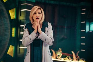 Jodie Whittaker as The Doctor, standing with her palms pressed together and a lock of shock on her face