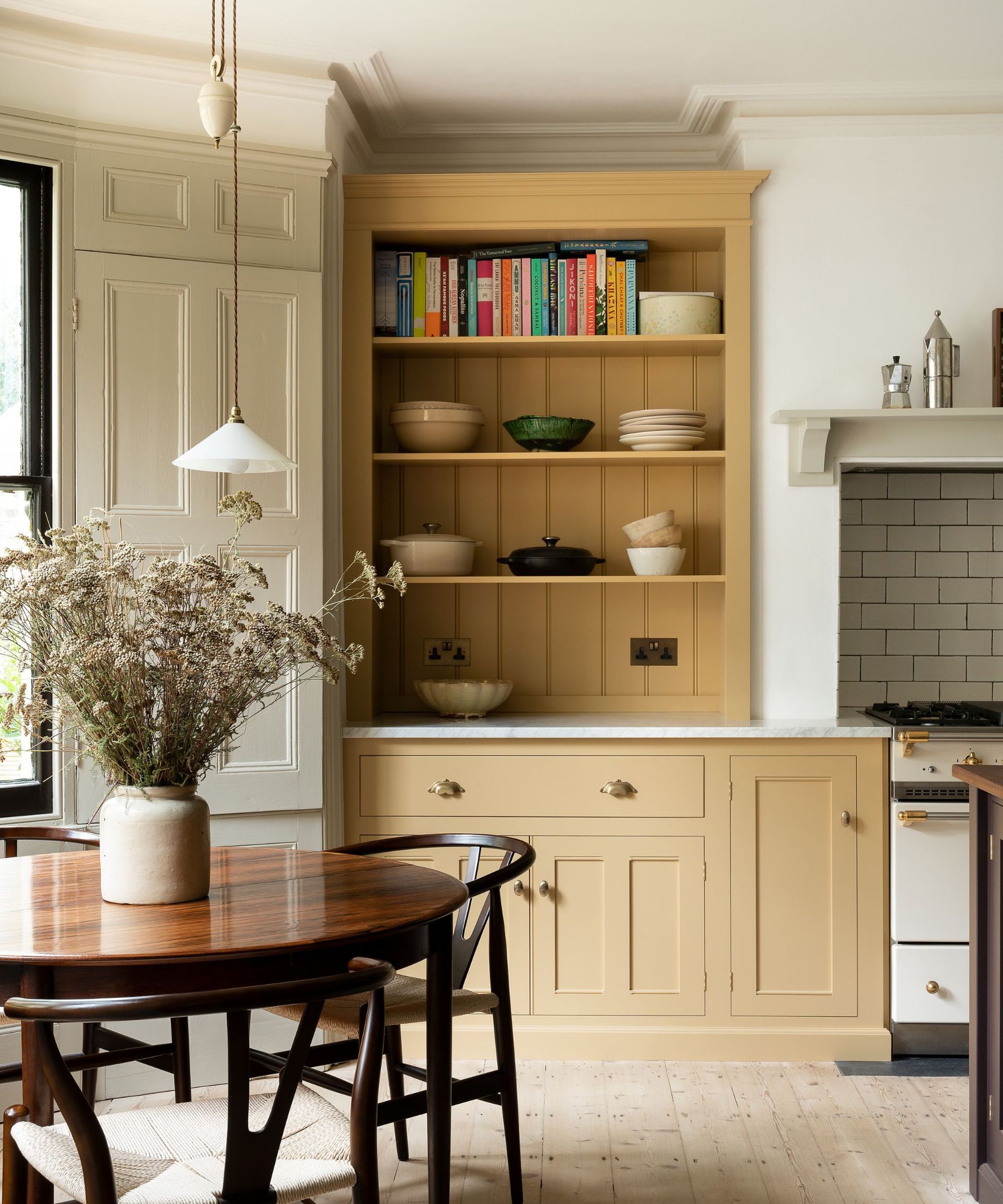 A kitchen with a large butter yellow built-in cabinet with open shelves. Colorful books lined up on the top shelf, with bowls and plates stacked on shelves below. A round dark wood dining table in front with matching curved chairs around it.