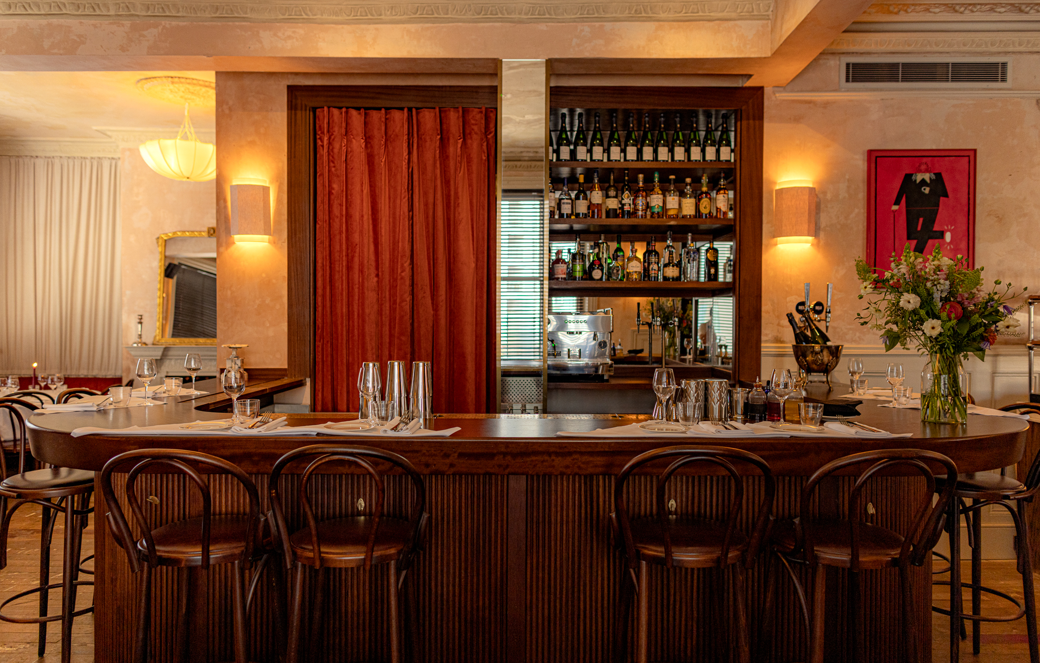 The charming-cool interiors of a French-style modern bistro with distressed plaster walls, lived-in wooden floors, elegant tables and chairs, colorful artworks, beige and red curtains, and sunset-hued sconces and pendant lights.