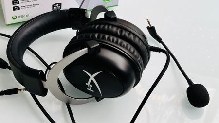 A black gaming headset with a detachable microphone and cushioned ear pads is displayed on a white surface. Xbox branding is partially visible in the background.