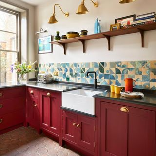 a kitchen countertop with dark red cabinets with brass cup pulls, and a striking blue graphic tiled splashback and. a large Belfast sink with black tap