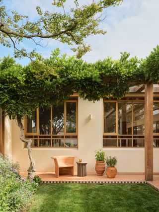 Neutral coloured exterior to the property and terracotta tile walkway with green lawn and plant pots in front. A pergola supports lush greenery at the full height of the windows and a tree sits to the left.