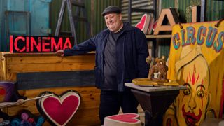 Johnny Vegas standing in a barn surrounded by antiques