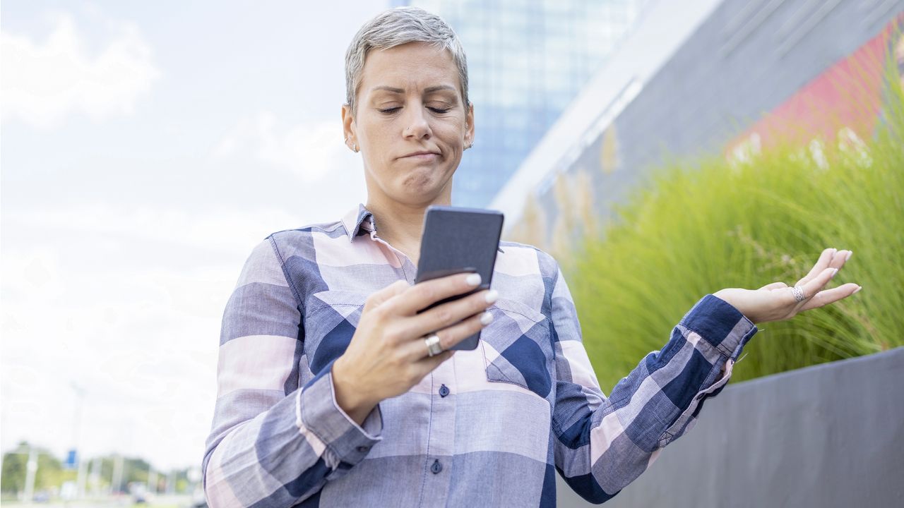 A woman who&#039;s walking outside gives her phone a frustrated look.