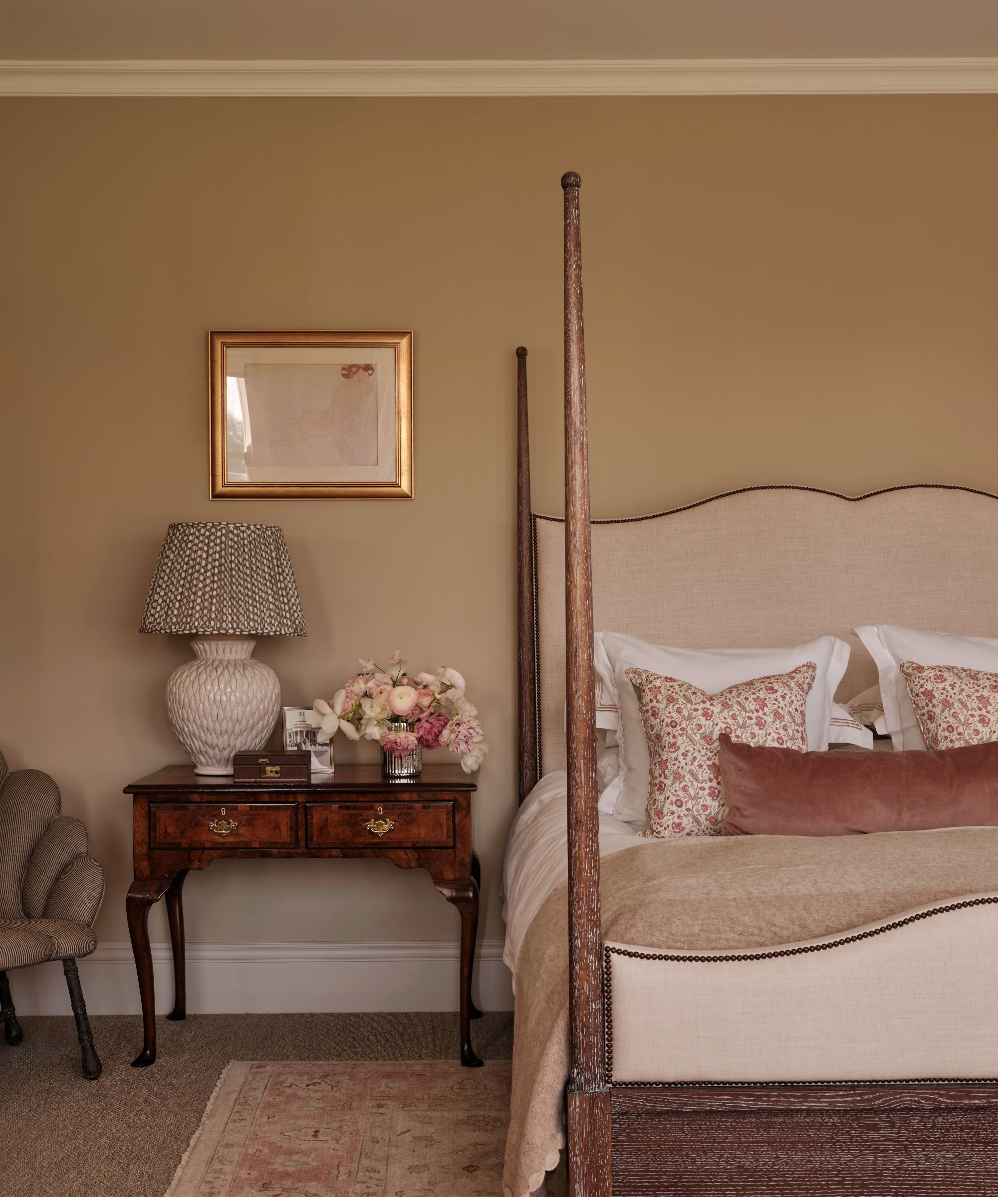 Bedroom with warm beige walls, pale pink upholstered four-poster bed with pale pink throw pillows and velvet sham, mahogany side table with pleated lap and flowers, and a pink Persian rug
