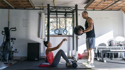 Fit Pacific Islander woman and muscular Caucasian man doing an abdominal strength workout with a weighted ball in a home gym set up in the garage of their home