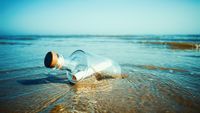 A large glass bottle with a cork holds a white rolled up piece of paper and sits on the beach.