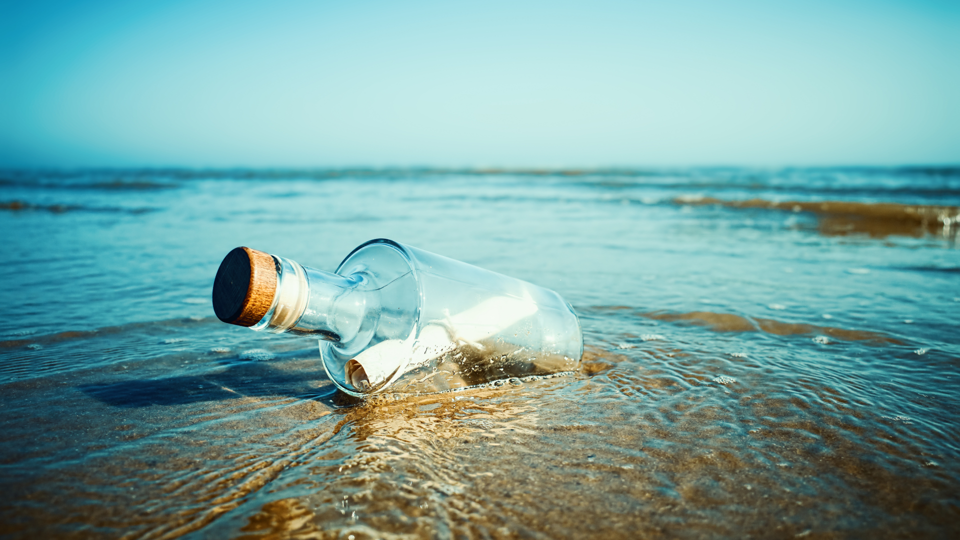 A large glass bottle with a cork holds a white rolled up piece of paper and sits on the beach.