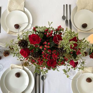 christmas flower arrangement containing red roses and pinecones sitting on a table with a white tablecloth and plates