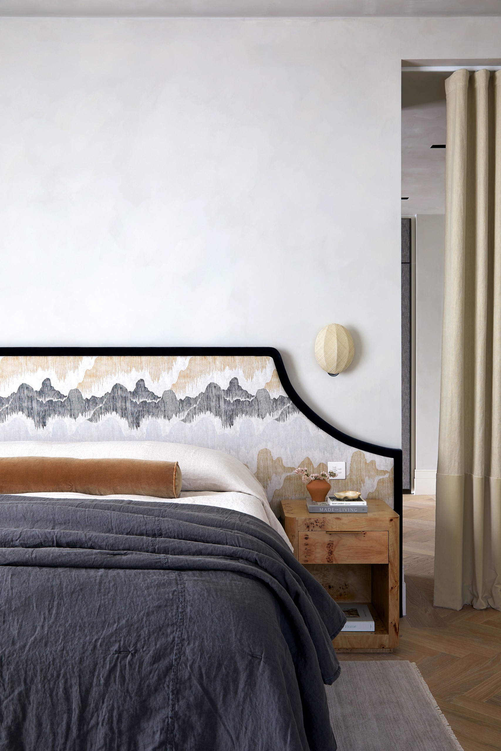 White bedroom with black-framed upholstered headboard with mountain print, charcoal bed sheets and terracotta bolster cushion, and burl wood coffee table