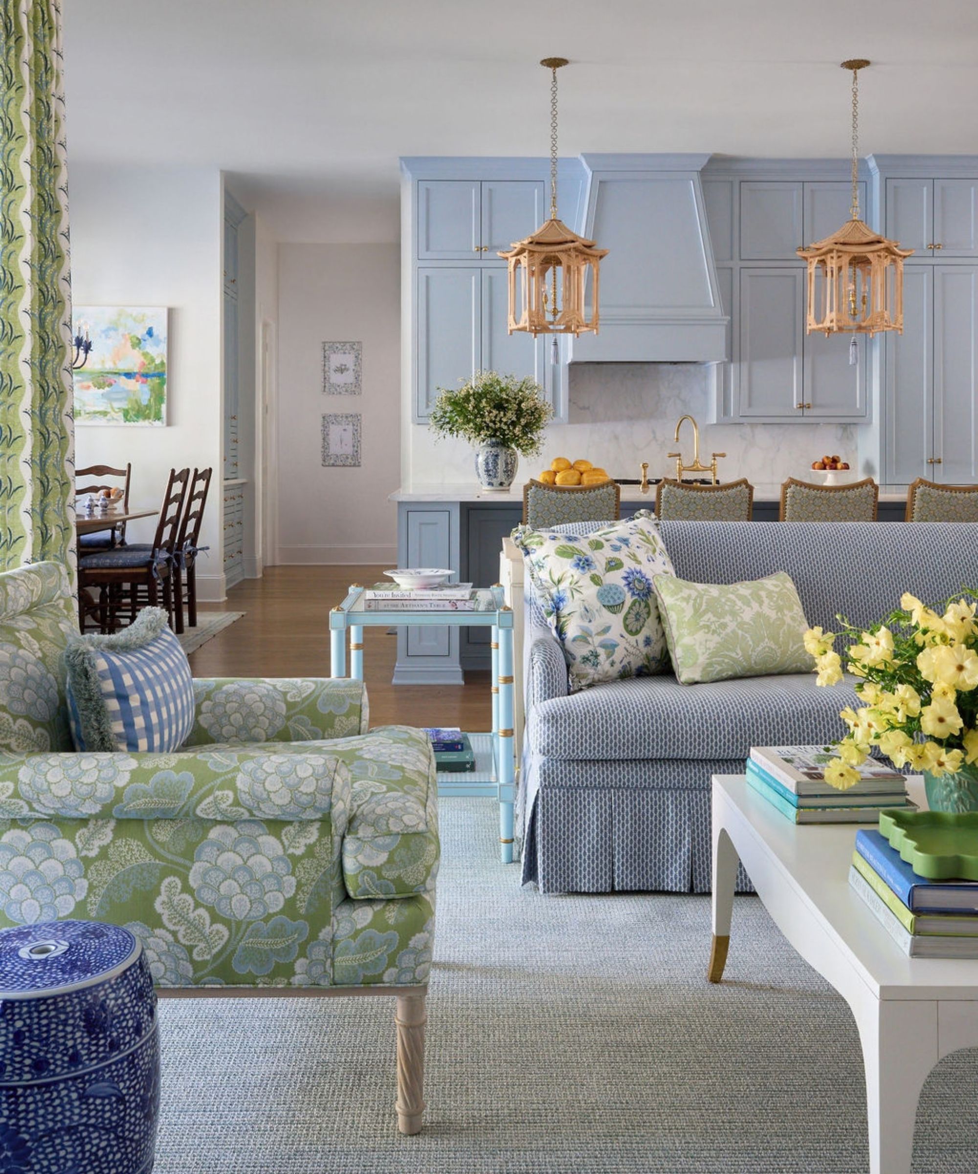 Open-concept living space showing a floral arm-chair and blue sofa in the foreground, leading into a light blue kitchen with pendant lights