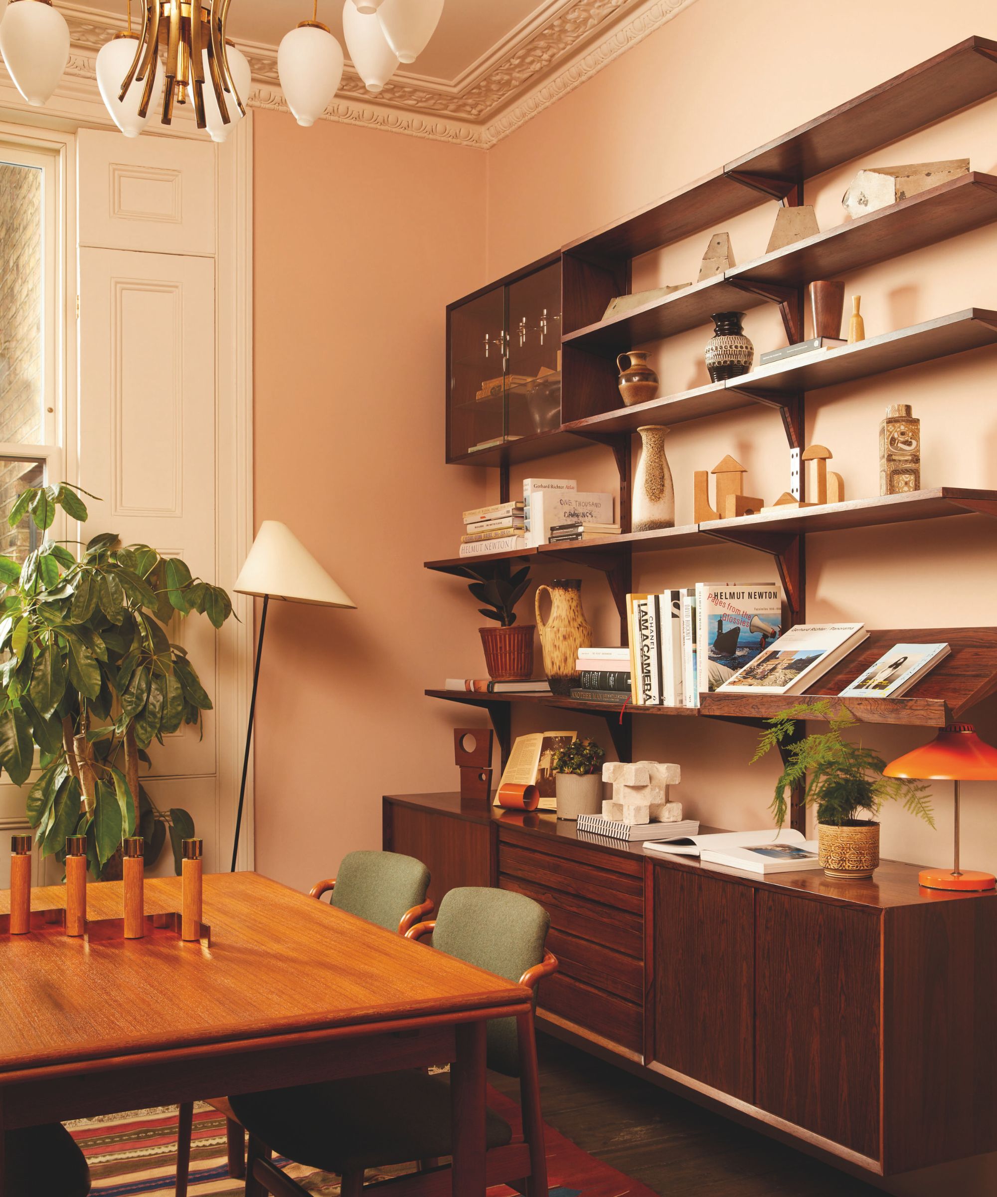 A warm-toned dining area with a mid-century modern teak wall unit filled with books and ceramics, set against soft terracotta-colored walls.