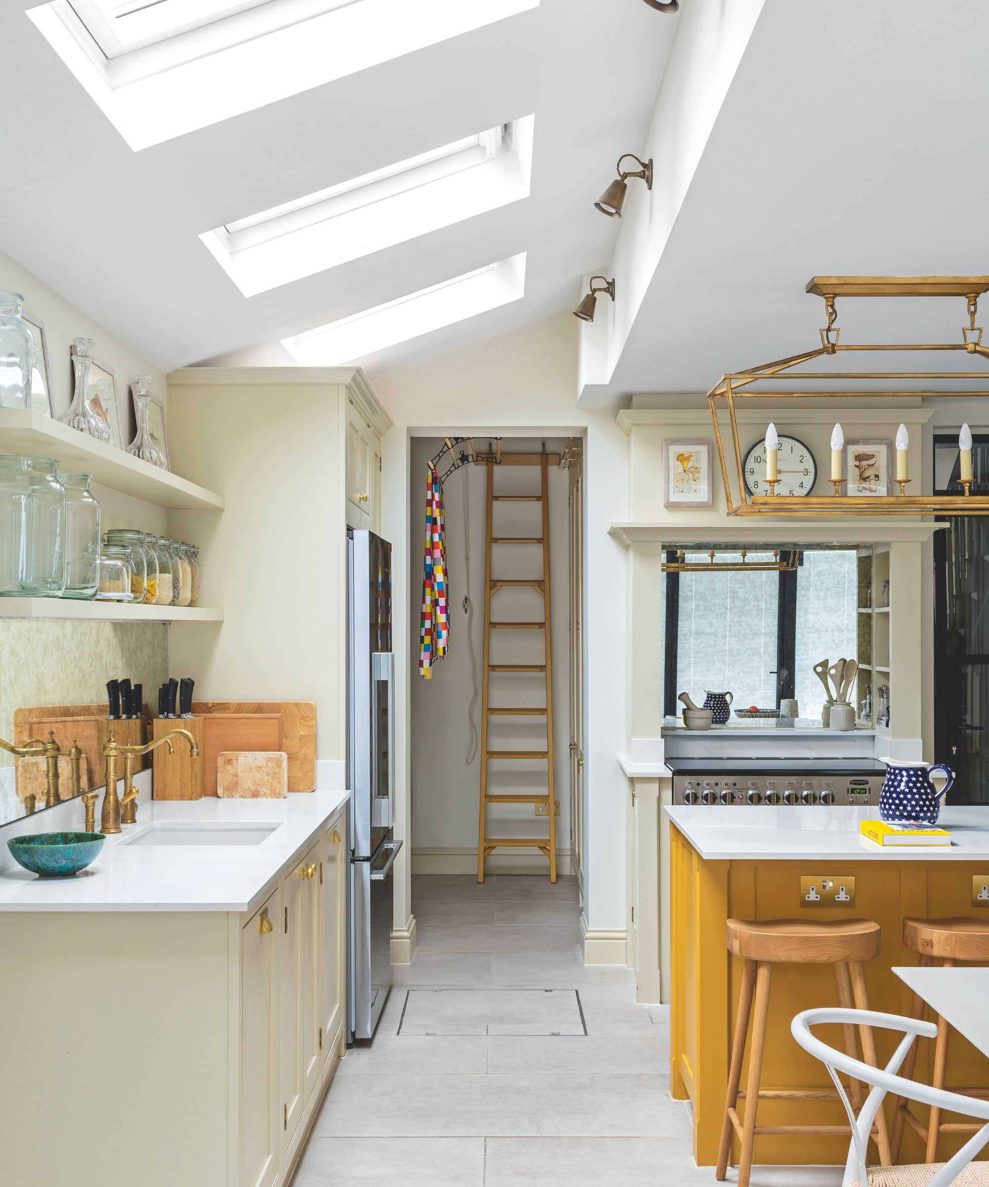 A bright kitchen with tiled floors, an ochre kitchen island, white countertops, wooden bar stools and skylights above.