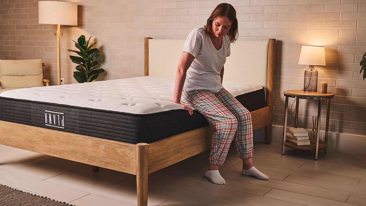 Our mattress tester Ruth Jones sits on the side of the Plank Firm mattress on a bed frame in bedroom. She is sitting comfortably on the edge, indicating good edge support