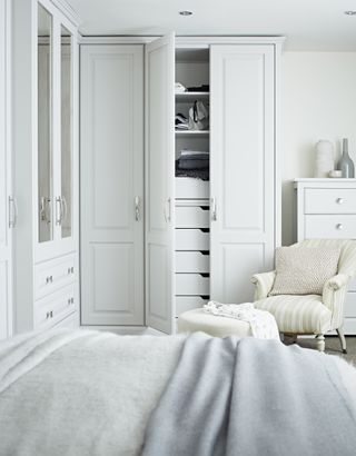 white bedroom with fitted closet doors in a corner, upholstered armchair, gray blankets, footstool, chest of drawers