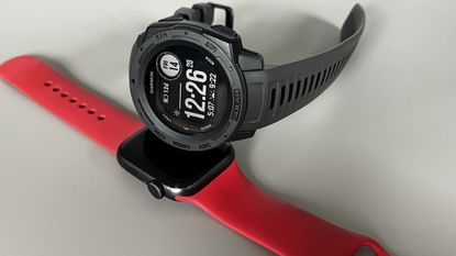 The Garmin Instinct Solar in Charcoal sat on top of an Apple Watch SE, on a grey background