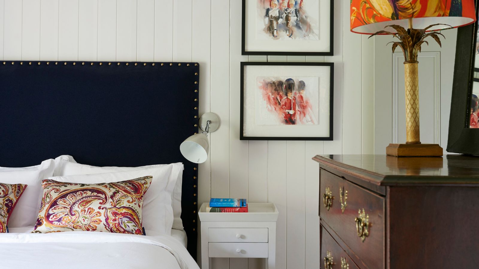 A white panelled bedroom with a double bed dark colored headboard, white bedding, patterned cushions, white nightstand, framed artworks, brown drawers, and statement lamp.