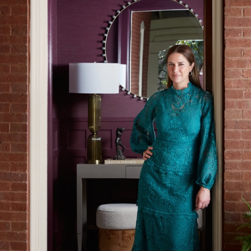 Image of a woman in a long-sleeve, teal dress leaning against a door frame with a purple entryway behind her.