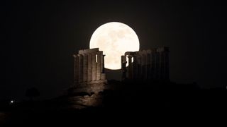 a large bright white moon appears behind the ruins of the Temple of Poseidon where several large columns rise high up into the sky. 